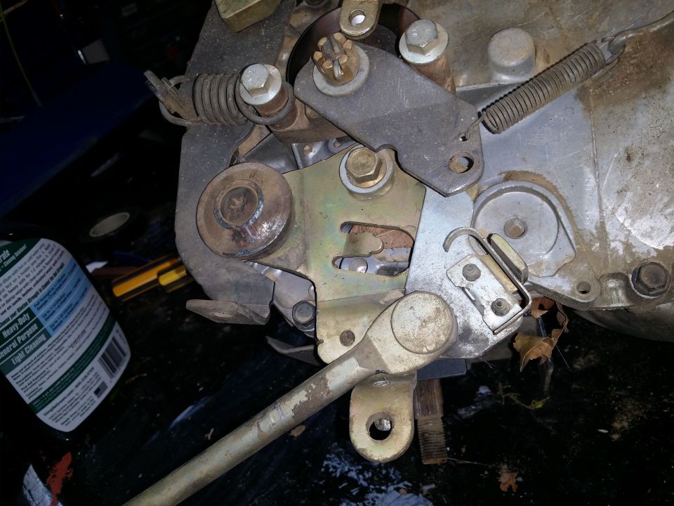 Close-up of a mechanical linkage assembly with springs, bolts, and levers. A bottle of cleaner is visible on the left