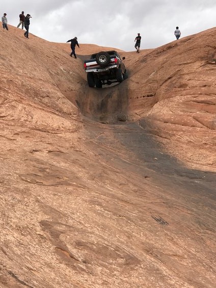 1991 Ford Bronco - A red and black SUV navigates a steep, rocky terrain with onlookers observing the challenging descent.