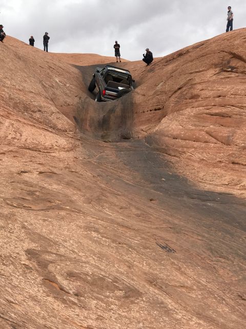 1991 Ford Bronco - An off-road vehicle is stuck in a precarious position on a steep, rocky incline while onlookers observe.