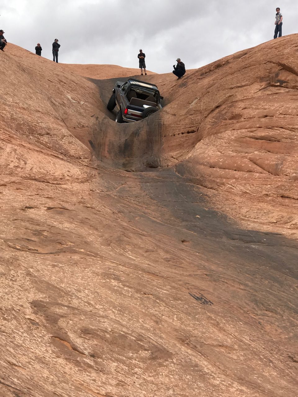 Dark SUV tilted in a steep, rocky ditch with people watching from above
