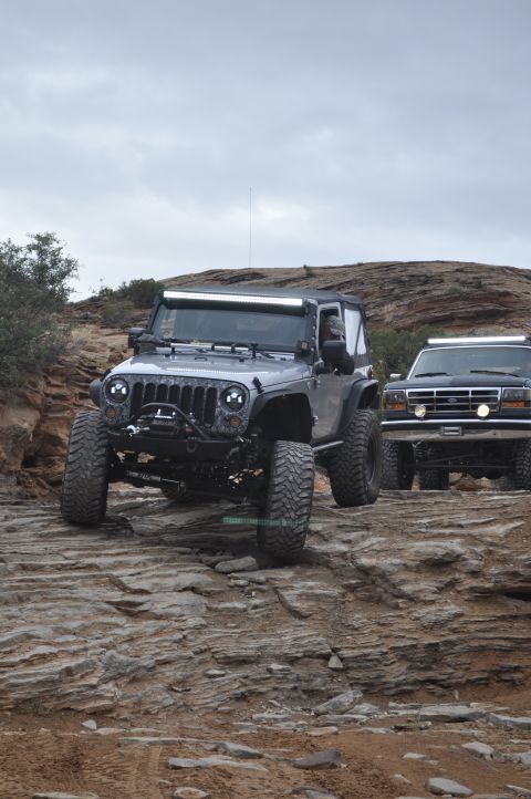 1991 Ford Bronco - Two modified SUVs, a Jeep Wrangler and a Ford Bronco, navigate a challenging rocky trail under an overcast sky.