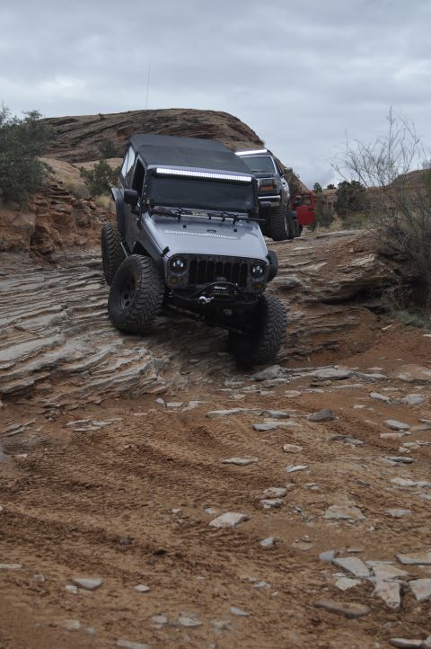 1991 Ford Bronco - Two Jeeps and a red vehicle navigate a challenging off-road trail with rocky terrain and a cloudy sky.
