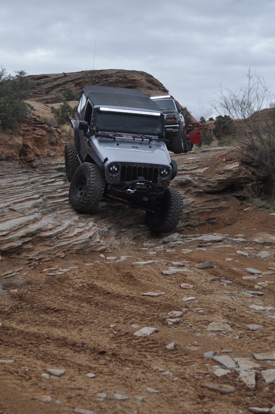 Silver Jeep Wrangler with a light bar driving up a rocky, off-road trail. Another Jeep and a red vehicle are behind it