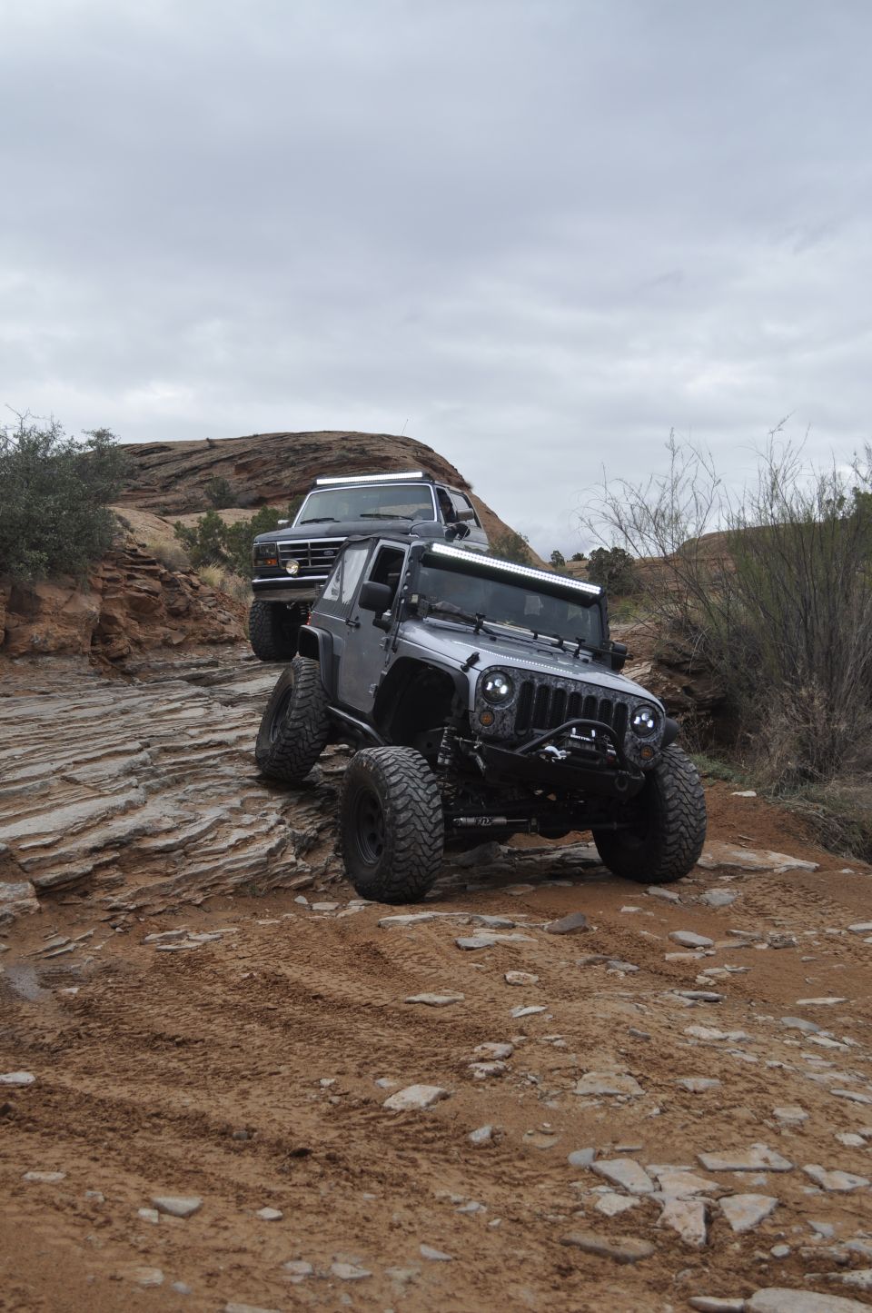 1991 Ford Bronco and a silver Jeep Wrangler off-roading on a rocky, dusty trail under a cloudy sky
