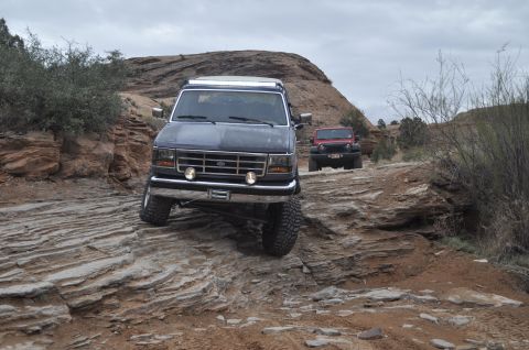 1991 Ford Bronco - A dark gray Ford Bronco with a roof rack drives on a challenging rocky trail, with a red Jeep following behind.