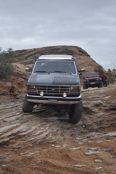 1991 Ford Bronco - A dark SUV navigates a challenging rocky trail in Moab, with another Jeep visible in the background.