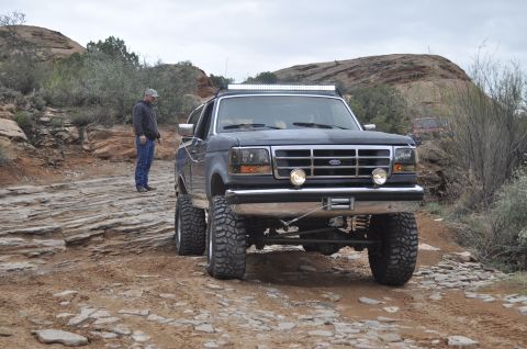 1991 Ford Bronco - A lifted Ford F-Series truck navigates a challenging, rocky terrain with a person observing the path ahead.