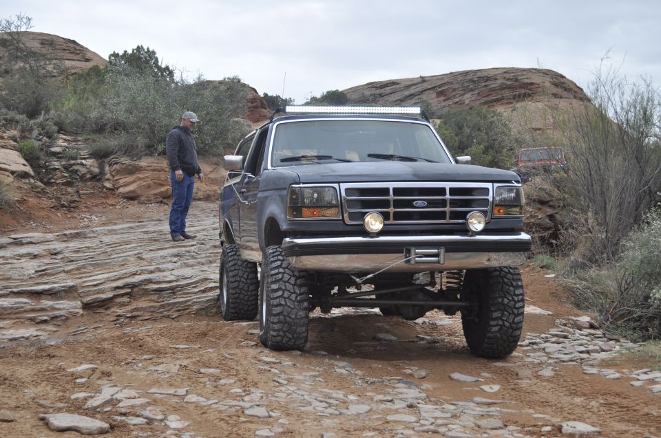 Ford F-Series truck on rocky off-road trail with a person standing nearby. Another vehicle is visible in the background