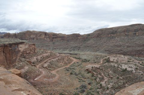 1991 Ford Bronco - A winding dirt road cuts through a desert canyon with layered rock formations and sparse vegetation.