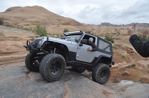 1991 Ford Bronco - A silver Jeep Wrangler tackles a challenging rocky trail, with a person giving a thumbs-up in the foreground.