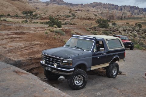 1991 Ford Bronco - Two off-road vehicles, a Ford Bronco and a Jeep Wrangler, navigate a rocky trail in a desert landscape.