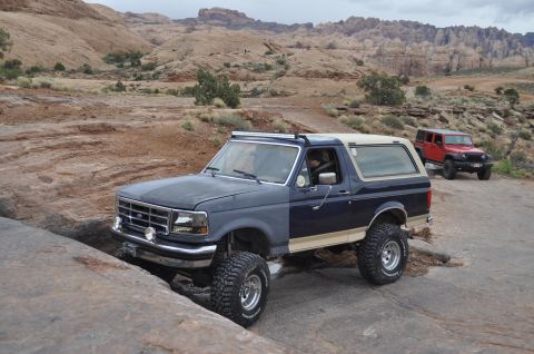 1991 Ford Bronco - Two off-road vehicles navigate a , rocky landscape under a cloudy sky.