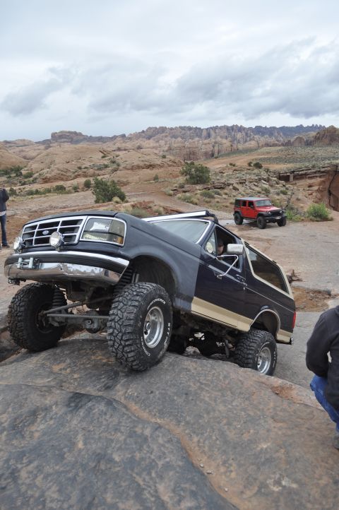 1991 Ford Bronco - A dark blue Ford Bronco with a tan stripe navigates rocky terrain, with a red Jeep in the background.