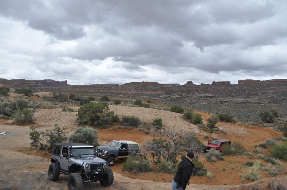 1991 Ford Bronco and two Jeeps on a dirt trail in a desert with rock formations under a cloudy sky