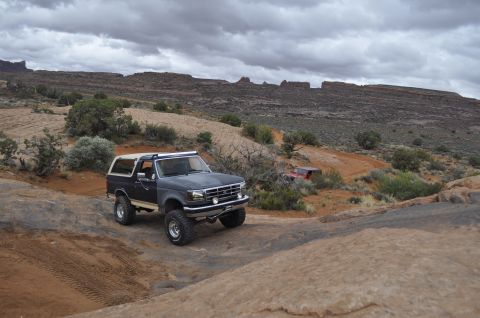 1991 Ford Bronco - A dark blue Ford Bronco navigates a rocky off-road trail under a cloudy sky.