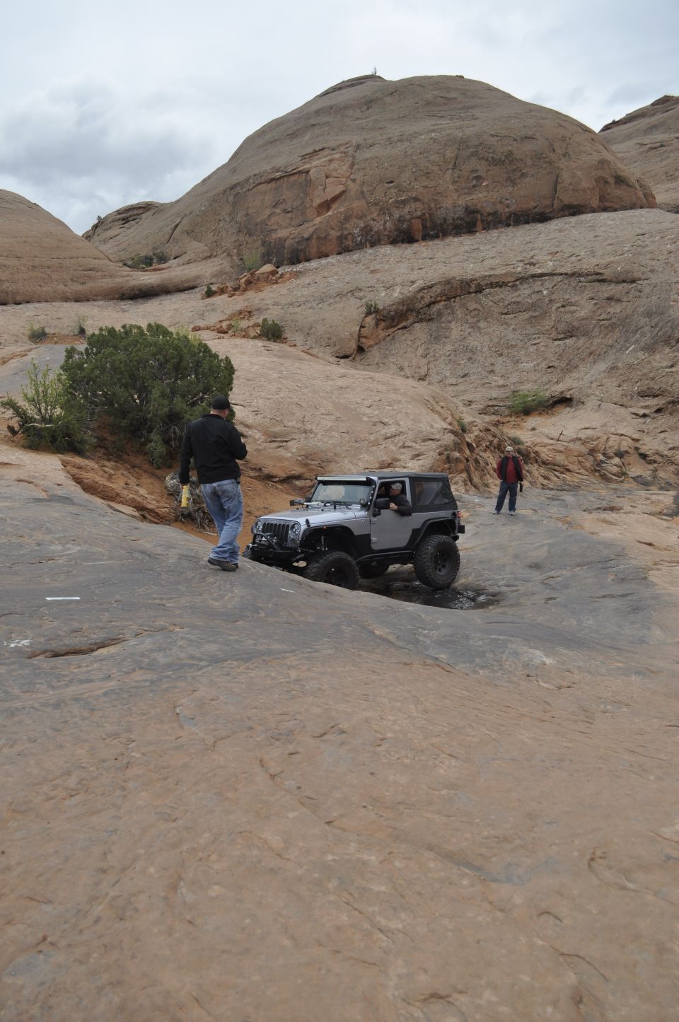 Silver Jeep Wrangler driving on rocky terrain with two people nearby