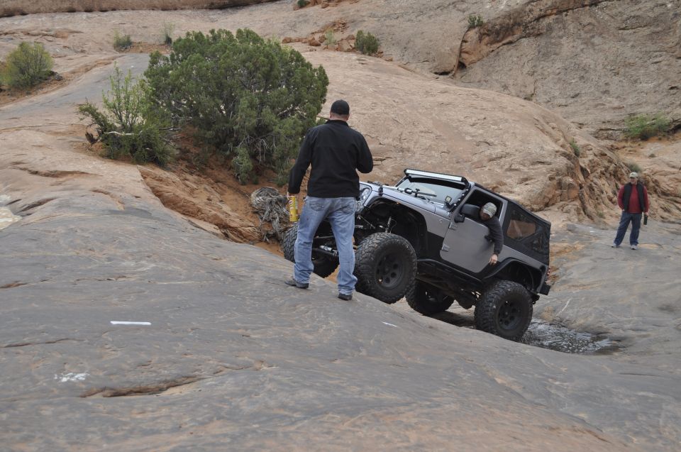 Silver Jeep Wrangler on rocky terrain, driver's door open, with a person standing nearby