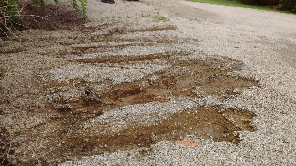 Eroded gravel driveway with deep washout ruts exposing brown dirt, bordered by brush