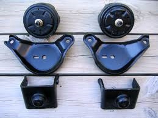 Four black automotive parts, likely engine mounts and brackets, arranged on a wooden surface