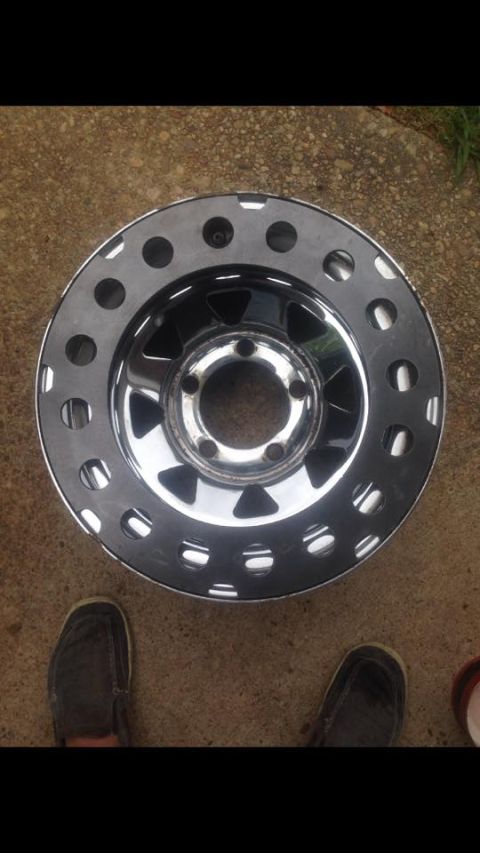1972 Ford Bronco - A single chrome wheel with a distinctive hole pattern sits on a concrete surface, with two sets of feet visible nearby.