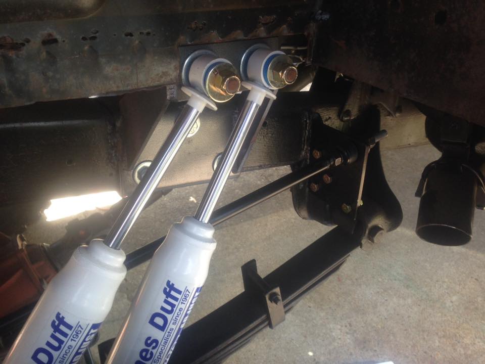 Close-up of two white shock absorbers with blue lettering, attached to a vehicle's rusty frame and leaf springs