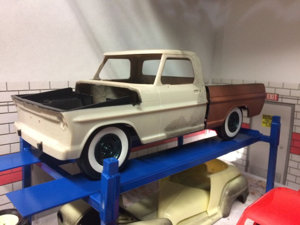 Model truck with cream cab and brown bed on a blue lift, another model car below