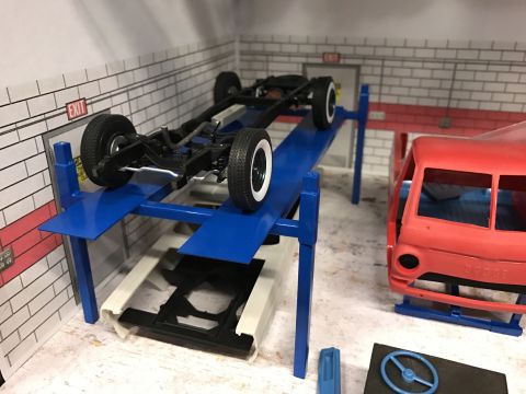 1989 Ford Bronco - A model car chassis is elevated on a repair lift, next to a red model truck body.