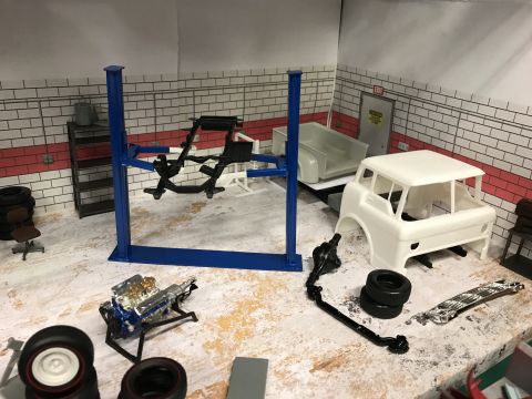 1989 Ford Bronco - A model garage scene displays a white car body, engine, tires, and a blue car lift on a workbench.