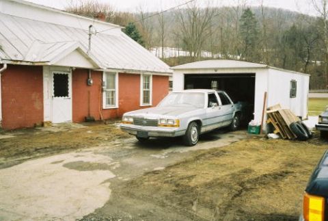 1989 Ford Crown Victoria LTD - A light blue sedan is parked in a driveway next to a red house and a white garage. Another vehicle is partially visible on the right.