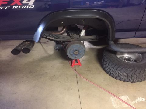 1982 Ford Bronco - The rear suspension and axle of a blue truck are visible, with the wheel removed and the vehicle supported by a jack stand.