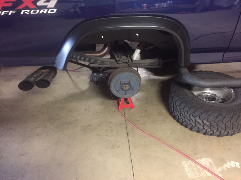 Blue truck's rear axle on a red jack stand, with dual exhaust pipes and a removed wheel