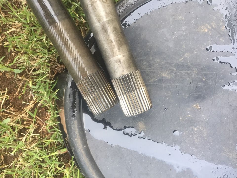 Two metal shafts with splined ends resting on a dark surface with water and grass in the background