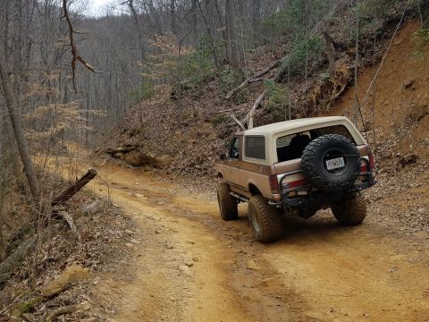 1988 Ford Bronco - A lifted Ford Bronco navigates a muddy trail through a forest.
