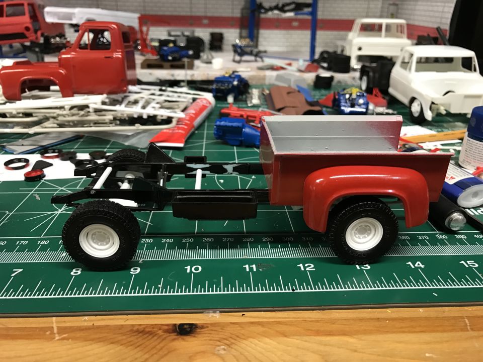 Model truck chassis with red cab and bed on green cutting mat, other model parts and vehicles in background