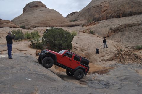 1991 Ford Bronco - A red Jeep Wrangler navigates a steep, rocky trail in a desert landscape with onlookers.