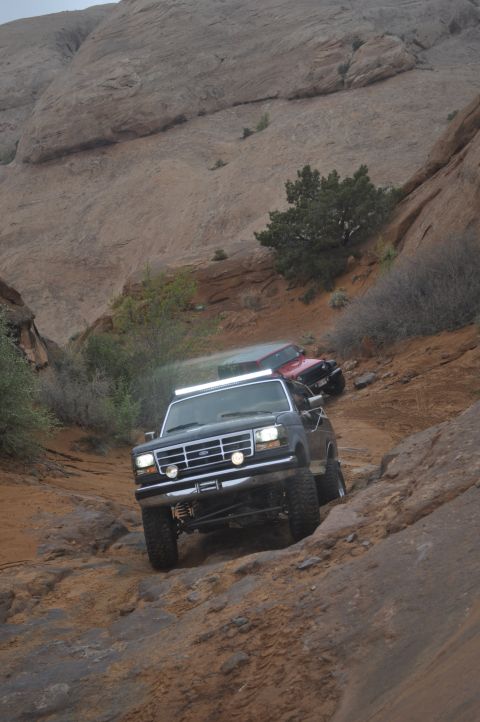 1991 Ford Bronco - Two SUVs navigate a steep, rocky off-road path in a desert canyon.