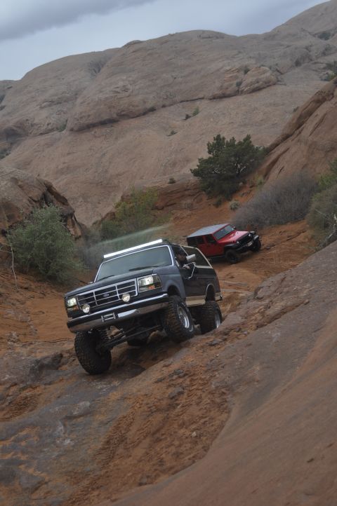 1991 Ford Bronco - Two off-road vehicles navigate a steep, rocky trail under a cloudy sky.