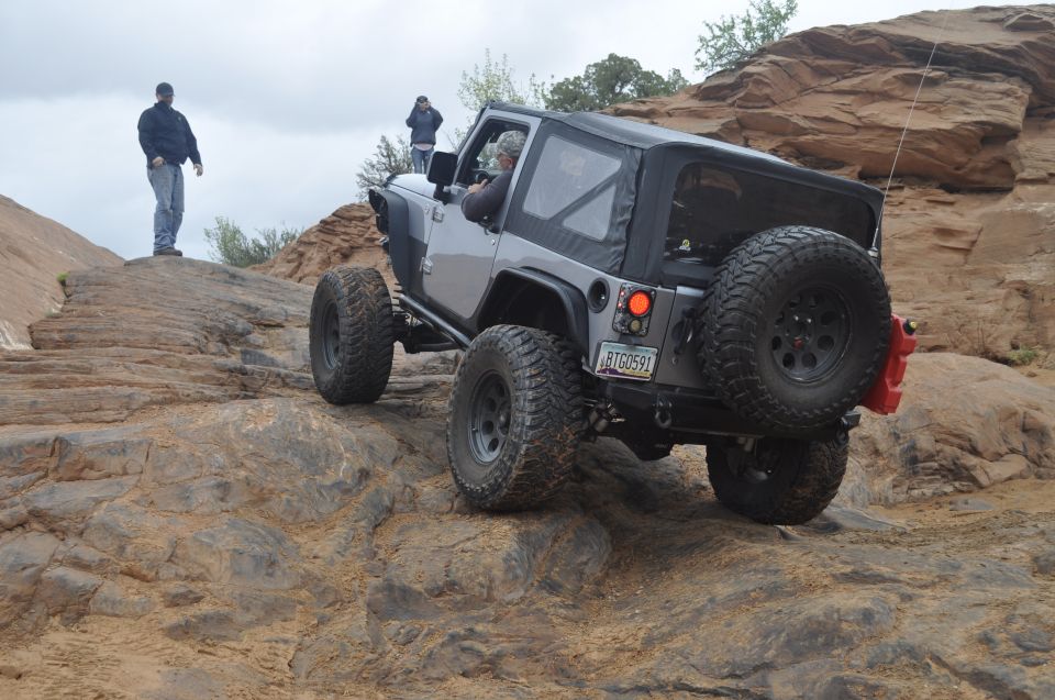 Silver Jeep Wrangler with a soft top navigating rocky terrain, rear view