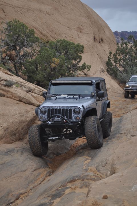 1991 Ford Bronco - A modified Jeep Wrangler tackles a challenging off-road trail in a rocky, desert landscape, with another vehicle following.