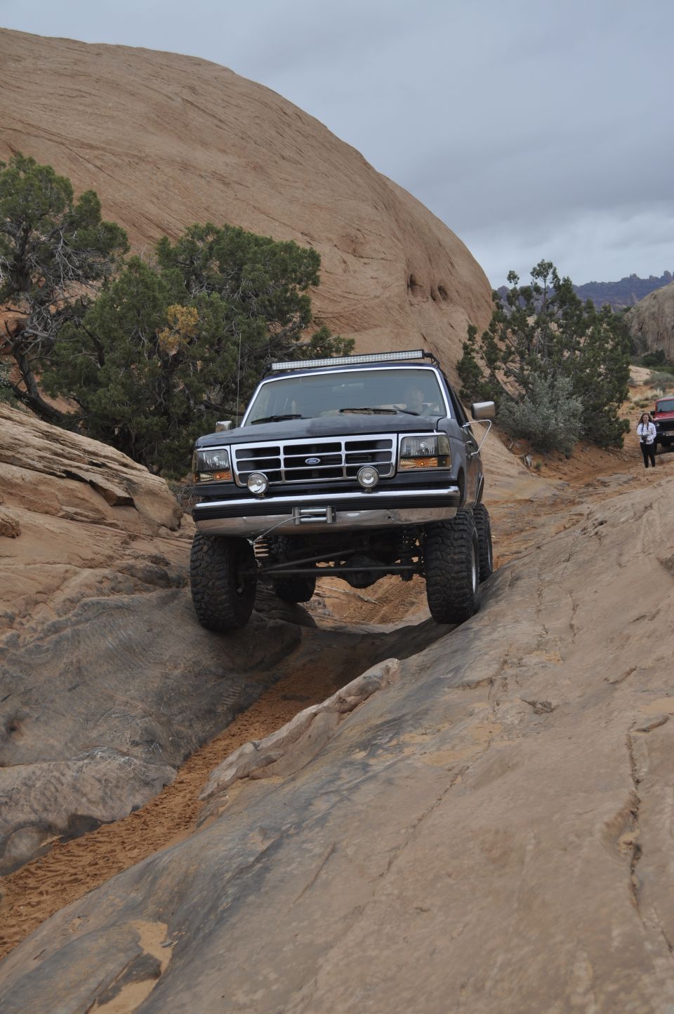 Dark blue Ford Bronco with light bar on roof climbing a rocky trail; another vehicle visible in background