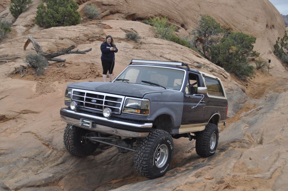 1991 Ford Bronco with lifted suspension and off-road tires navigating a rocky trail, a person stands in the background
