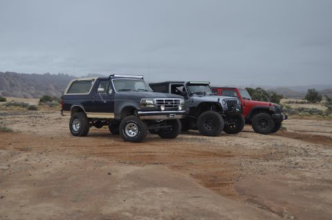 1991 Ford Bronco - Three modified off-road vehicles are parked in a dusty, arid landscape under an overcast sky.