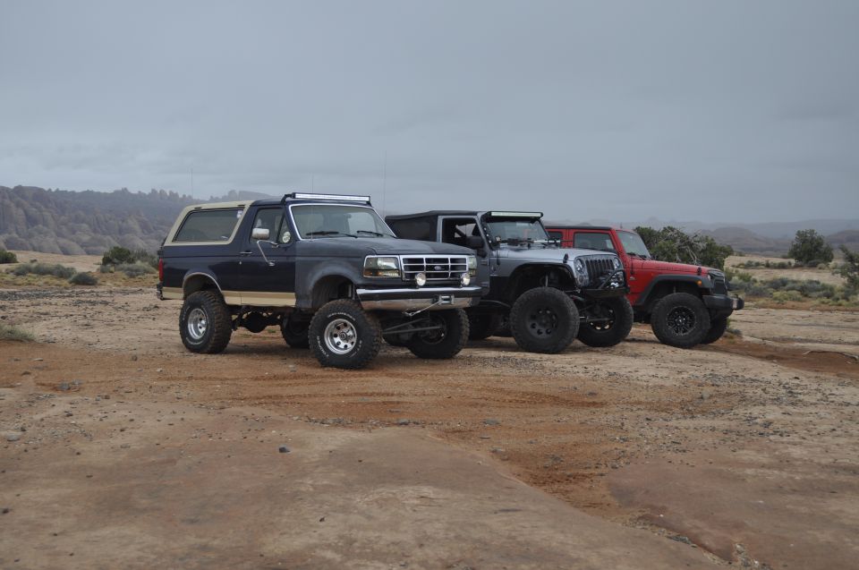 Dark blue Ford Bronco, silver Jeep Wrangler, and red Jeep Wrangler parked on dirt with hills in background