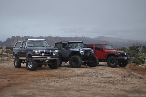 1991 Ford Bronco - Three modified off-road vehicles are parked in a desert landscape under a cloudy sky.