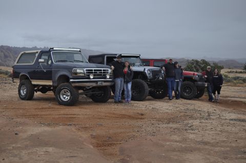 1991 Ford Bronco - A group poses with their lifted off-road vehicles, including a dark blue 1991 Ford Bronco, in a desert landscape.