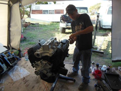 1984 Ford Bronco - Man works on a V8 engine assembly on a stand, with other engine components nearby.