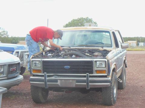 1984 Ford Bronco - A man works on the engine of a blue Ford Bronco, parked next to other vehicles.
