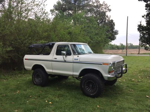 1979 Ford Bronco - This lifted white Ford Bronco sits on a grassy area, with trees and a utility pole visible in the background.