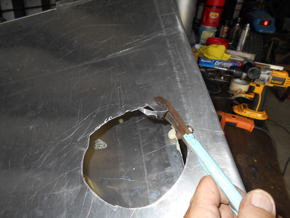 Tin snips cutting a circular hole in a large, scratched metal panel. Tools are visible in the background