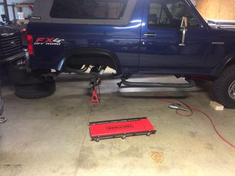 1982 Ford Bronco - A blue Ford Bronco is parked in a garage, lifted on a red jack stand with its rear axle exposed. A red creeper sits on the floor in front o…